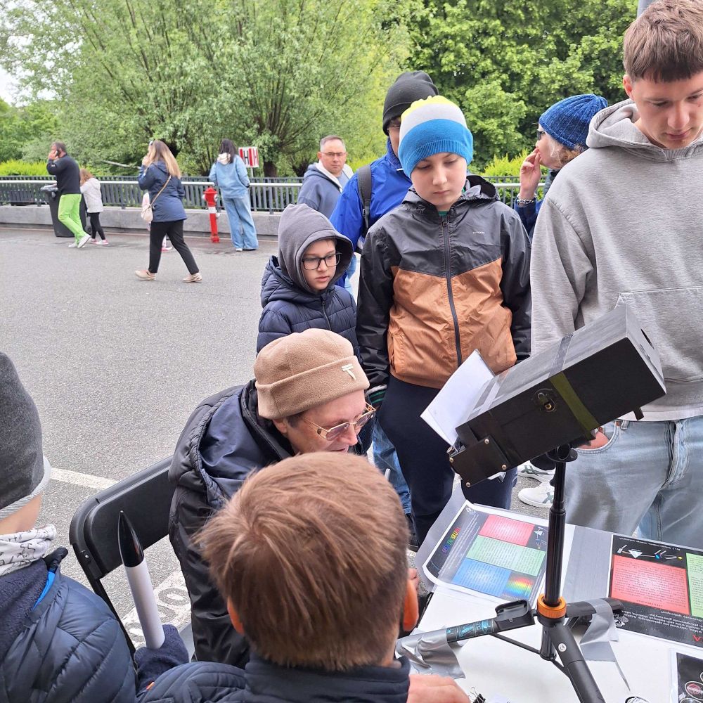 Outdoors a volunteer demonstrates a black-boxed telescope-style device on a tripod to a group of curious children, with colourful spectra charts spread on the table.