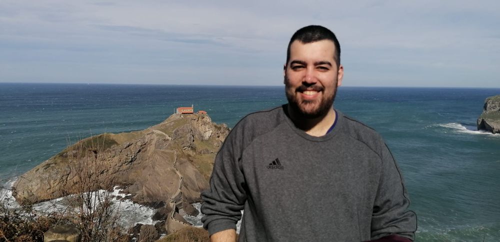 A person smiling in front of the ocean, with Gaztelugatxe — a rocky island featuring a small chapel — visible in the background on a sunny day