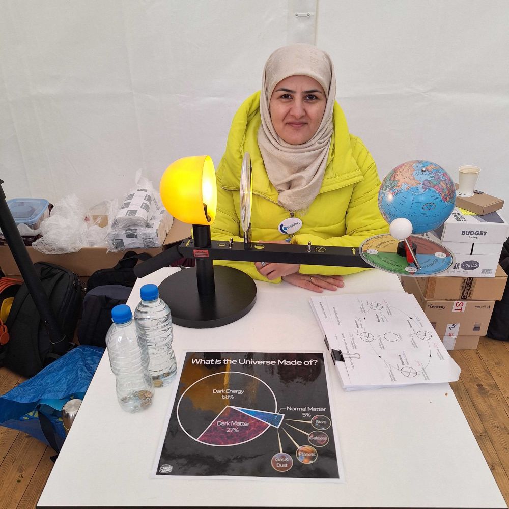 A presenter in a yellow coat and beige headscarf sits behind a Sun–Earth–Moon demonstration model; on the table is a poster titled “What is the Universe Made of?” highlighting dark matter and dark energy.
