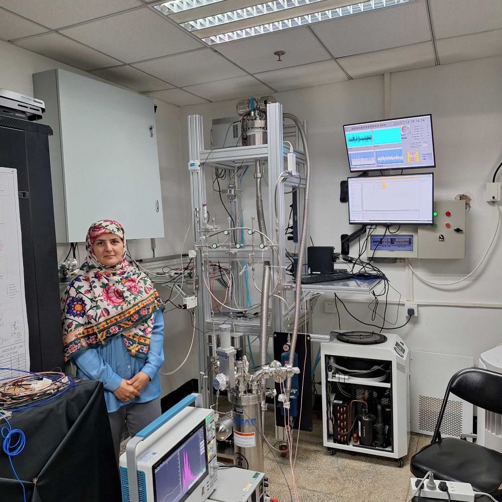 A researcher in a floral headscarf stands beside a tall cryogenic detector system with multiple cables and control sensors. Computer monitors in the background display real-time data charts and graphs.