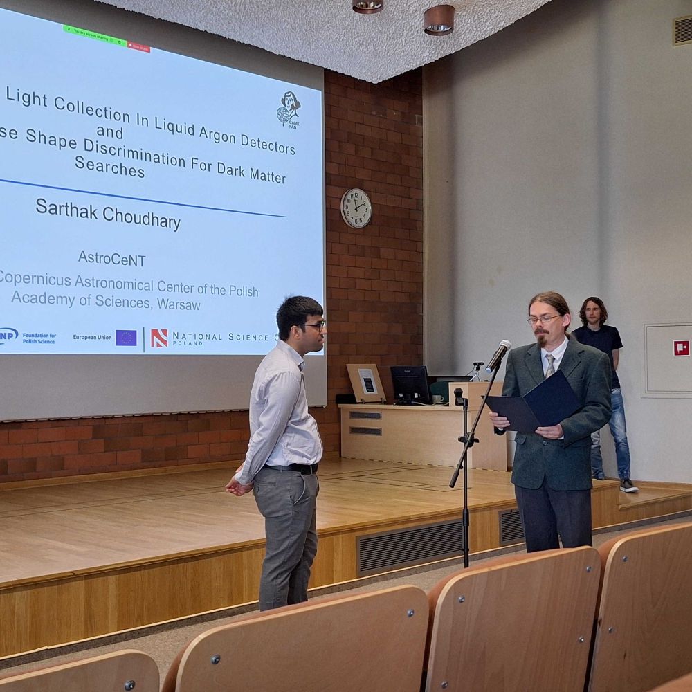 The PhD candidate stands on stage in front of the screen showing the thesis title, listening to a faculty member delivering final remarks at a microphone. The atmosphere is formal, with a few people visible in the background.