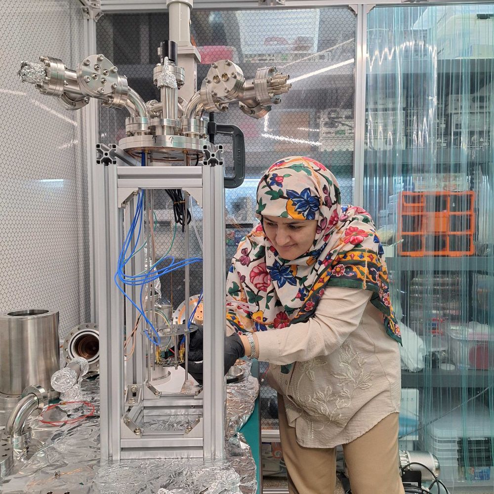 A researcher wearing a colorful floral headscarf and light clothing is working on an experimental cryogenic detector system with aluminum foil-wrapped components and connected blue wires. Laboratory shelves with tools and containers are visible in the background.