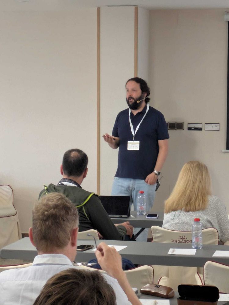 A speaker giving an invited talk at the IBER 2025 conference in Vigo, Spain. The person is standing near a screen displaying a presentation titled “Enhancing light collection of next-generation Dark Matter detectors – from wavelength-shifters to novel hybrid optical structures,” with the Astrocent logo visible.