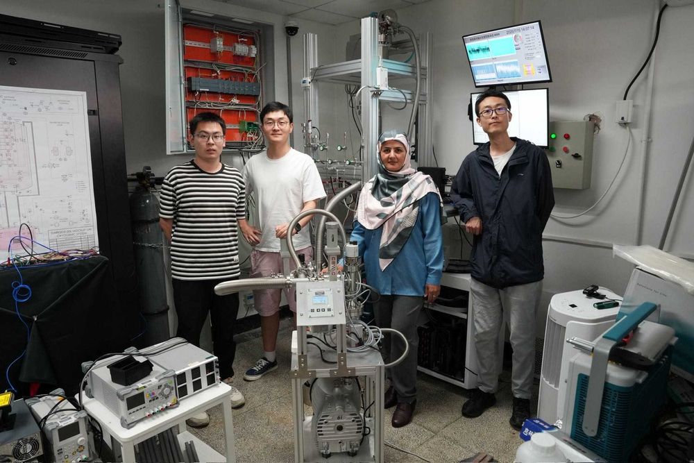 Four researchers stand in a laboratory next to a complex cryogenic and detector setup with stainless steel tubes, cables, and sensors. Behind them is an open control cabinet with wiring, monitors showing data plots, and electronic equipment on tables.