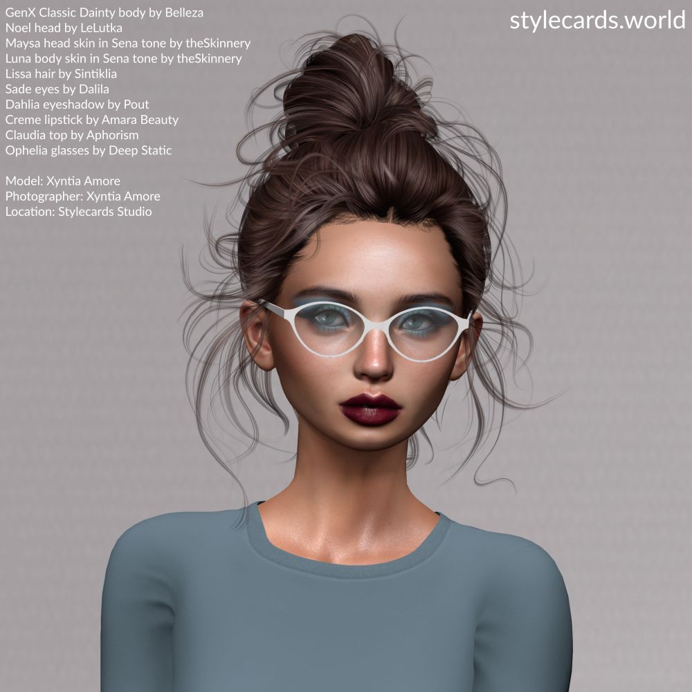 Stylecards Editor Xyntia Amore poses in front of a grey backdrop. She is wearing a slate blue long-sleeved t-shirt, has burgundy lipstick and pale blue eyeshadow. Her hair is done up in a bun, but is quite messy indicating her hard work. She is wearing a pair of white-framed eyeglasses.
