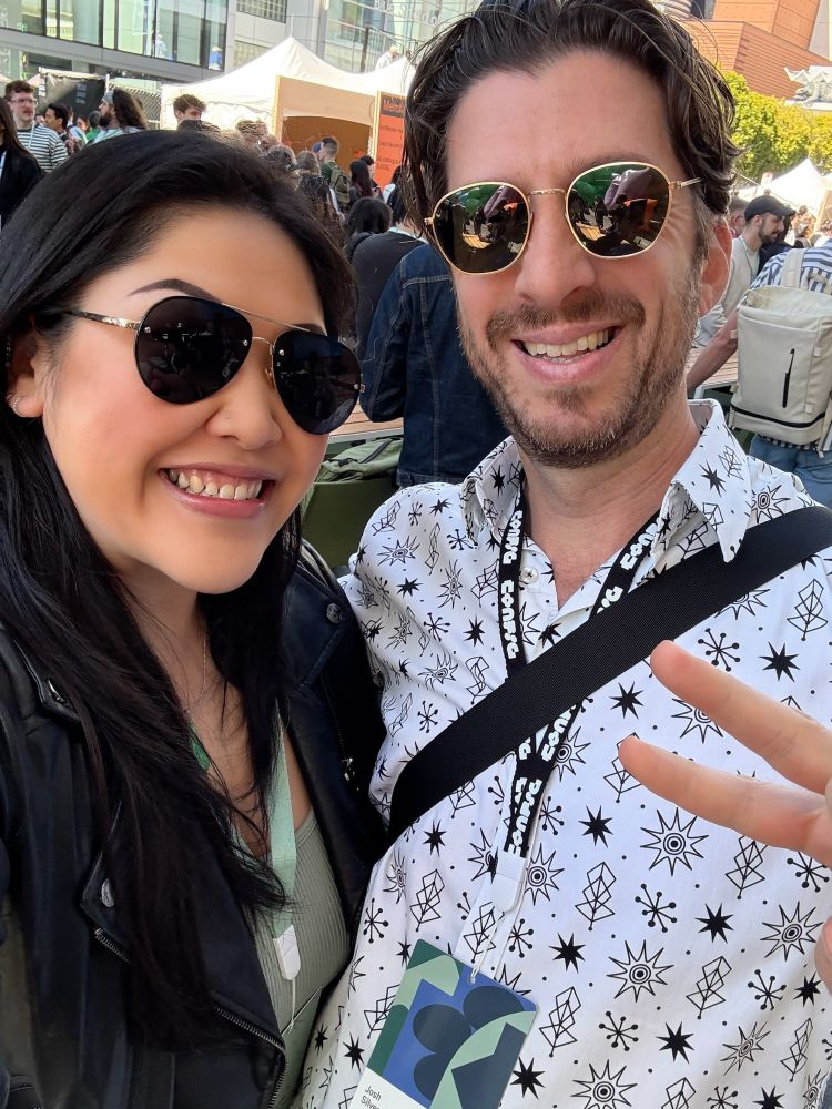 Photo of a woman wearing a black leather jacket, sage green cropped top, and sunglasses with a man wearing a white button up shirt with black patterns all over it and sunglasses while giving the peace sign