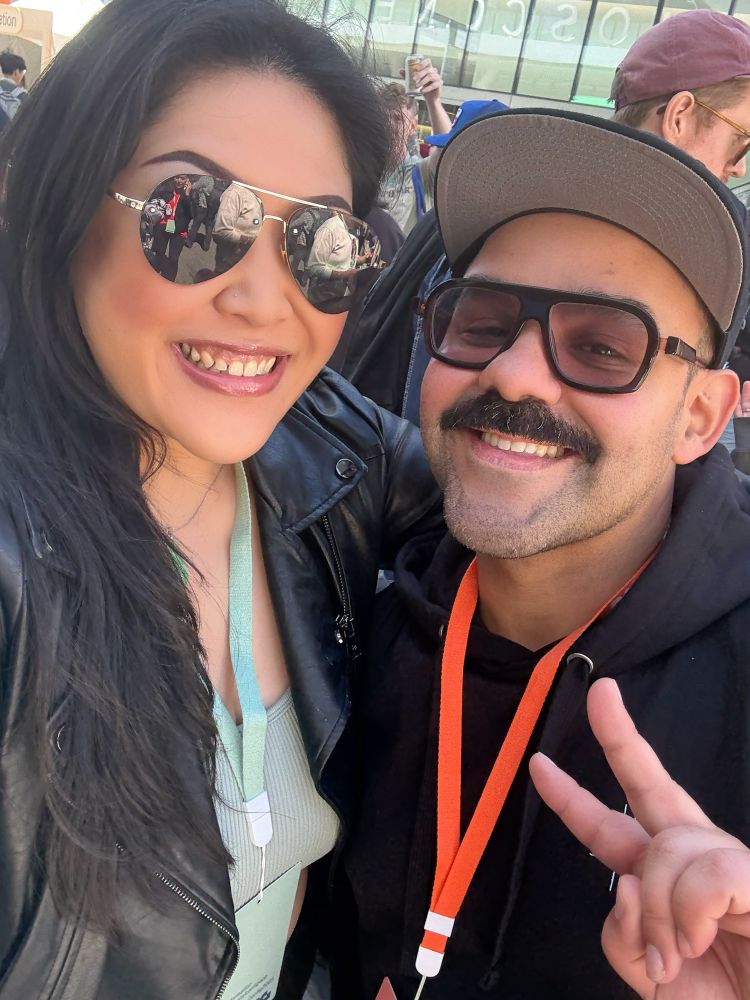 Photo of a woman wearing a black leather jacket, sunglasses, and a sage green cropped top next to a man wearing a black hoodie and sunglasses giving a peace sign with that super fresh mustache 