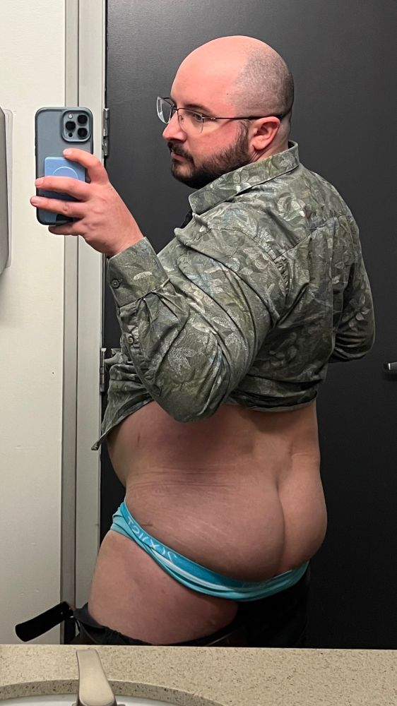 A man wearing blue underwear in a bathroom taking a selfie. The blue underwear is pulled down to view cheeks