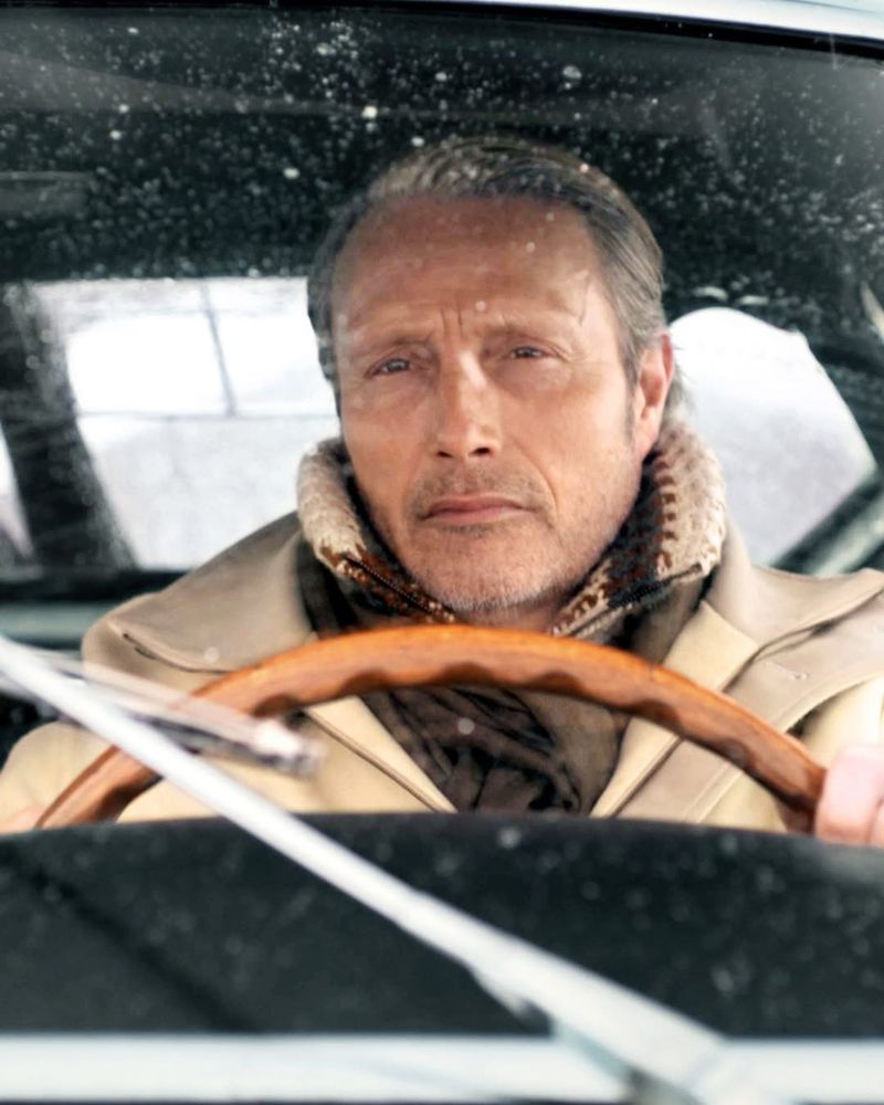 Mads Mikkelsen in an ad for Zegna. He’s wearing a khaki colored jacket over a high neck sweater, behind the drivers wheel in a car. 
