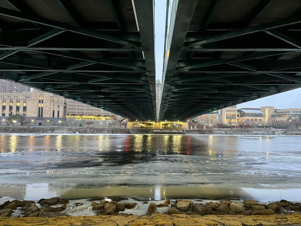 Underneath a bridge, half frozen river, yellow light reflected on the water.