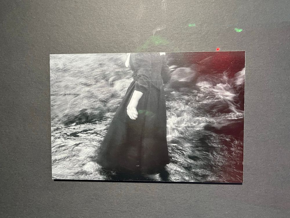 This is a black and white photograph by Joanne Coates of a figure in a dark dress making her way through the landscape. It is a close up, with the composition cutting off the head, to show the figure in movement. ‘Trajicere’, 2024, Joanne Coates 