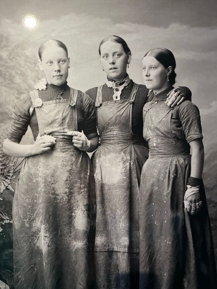 Three women wearing long aprons pose for a formal photograph. All have their hair pulled back tight. ‘Three women herring gutters July 1884’, Wick Society, Johnston Photographic Collection.