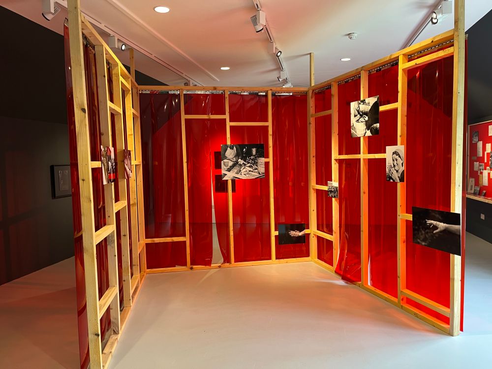 This wall construction is at the heart of the exhibition at Timespan. It is freestanding, with wooden frame visible. It has been covered on the outer side in a thick red plastic. Archival photographs and images are pinned to the interior surface of the framework