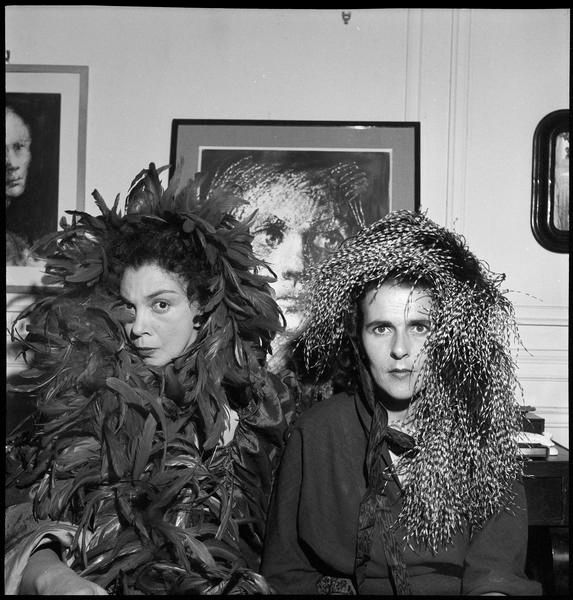 The two pose in front of a painting, almost completely covered in feathered headdresses.