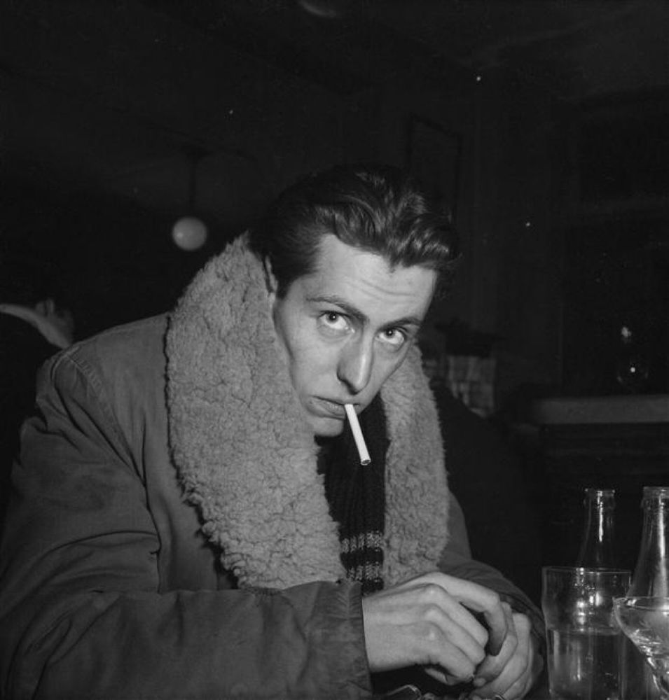 Buffet in a heavy sheepskin coat, lighting a cigarette in a Paris bar. 