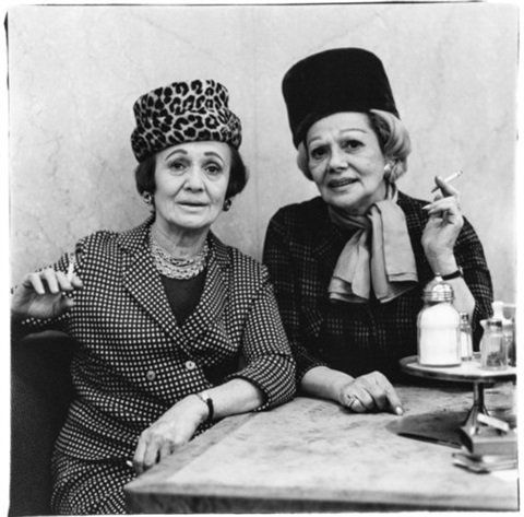 Two women at an automat table, both wearing hats, both smoking cigarettes.