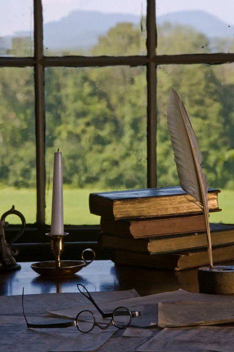 A stack of books, a feather quill, a candlestick & Melville's glasses, all on an old wooden desk, looking out a window to a hilly landscape.