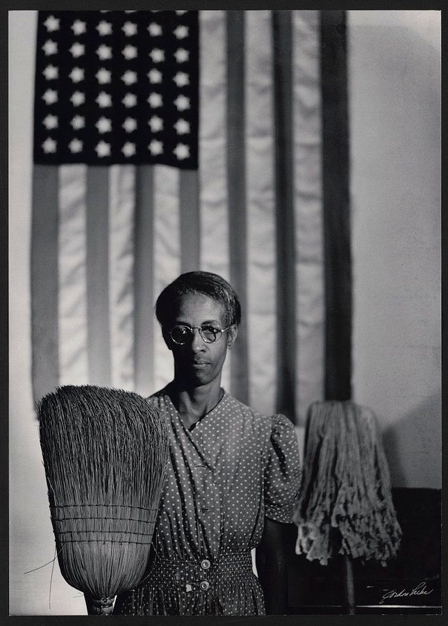 A woman poses with her broom & mop, in front of an American flag.