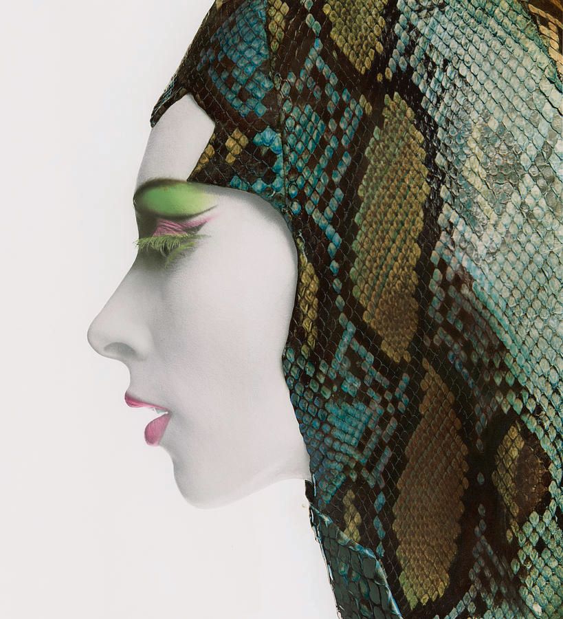 Description: Close-up of model in profile wearing white foundation makeup, green upside-down false eyelashes, purple and green eye shadow, and mauve lipstick all applied by Revlon's Stan Lawrence. Covering her head and surrounding her face is a multicolored snakeskin hood by Adolfo.