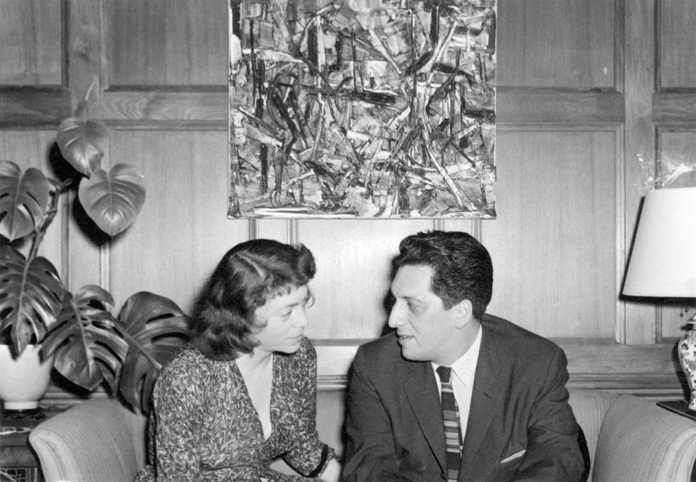 Mitchell & Riopelle sit on a sofa in front of an abstract expressionist painting