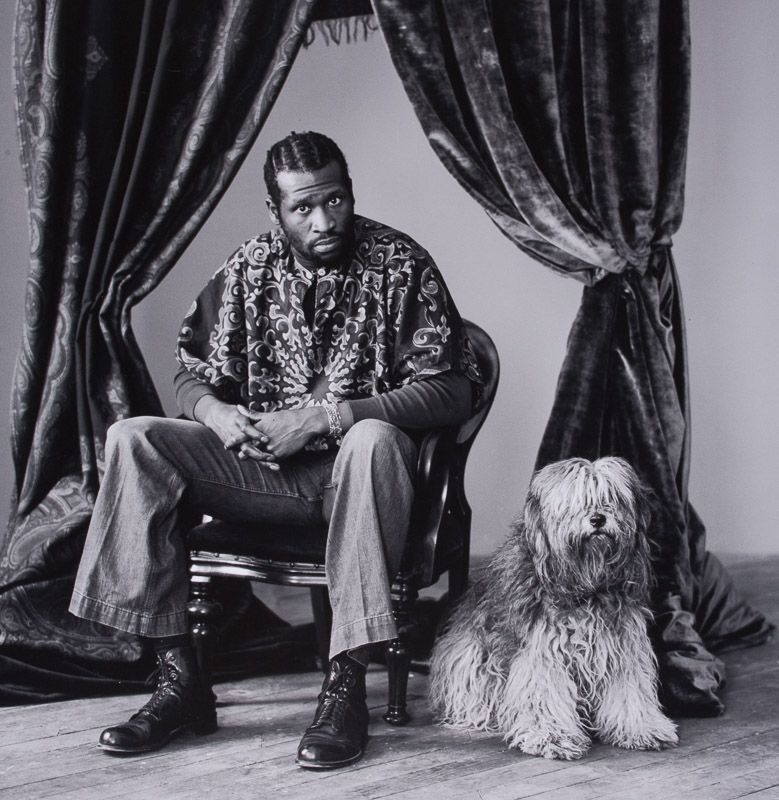 The musician sits on a chair, next to a sheepdog