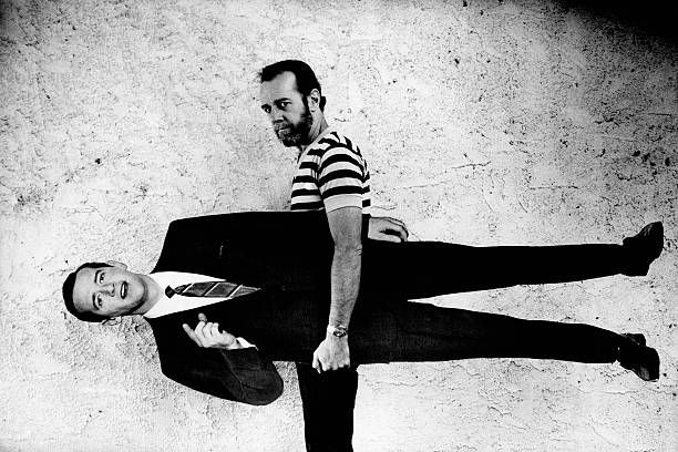 A bearded Carlin in a striped t-shirt carries a life-size cardboard figure of a clean-shaven, younger Carlin in a suit & tie.
