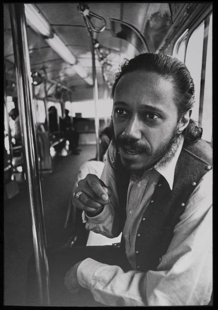 Wilmer smokes a cigarette on a bus. He's wearing a leather vest.