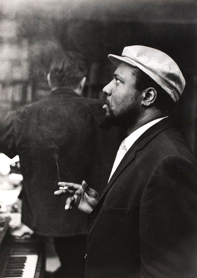 Monk in profile at the piano. He's wearing a soft cap & smoking a cigarette.