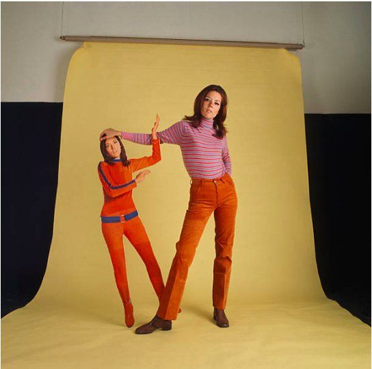 Rigg holds up a slightly smaller than lifesize cardboard cutout of Mrs. Peel, against a yellow background.