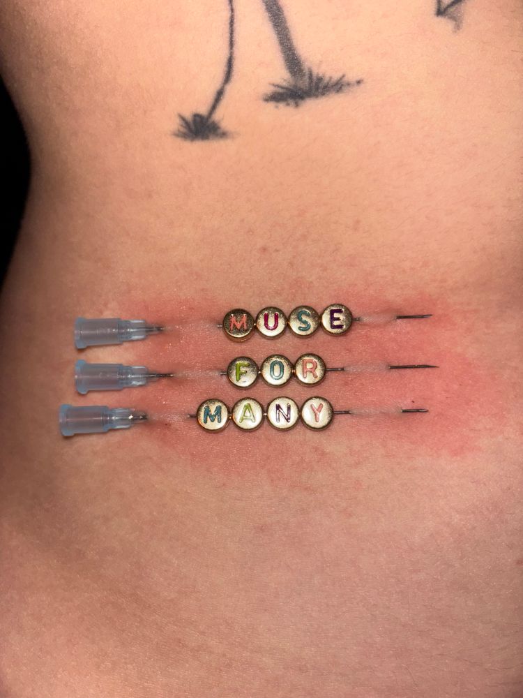 three long needles are pierced into flesh. the needles have beads threaded in between pierced flesh. the beads read “muse for many,” one word on each needle 