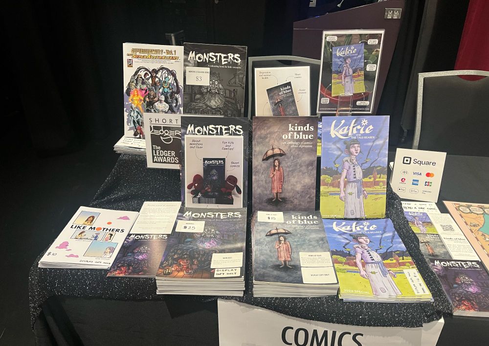 The comics half of my table, featuring print copies of “Katrie and the Tale-Bearer”, “Monsters”, “Kinds of Blue” and “Like Mothers”.