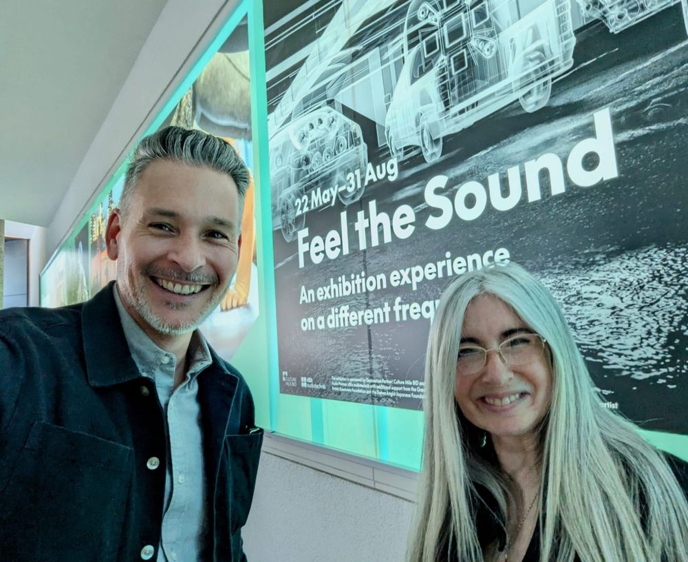 Gethyn Williams, Chair of The Evelyn Glennie Foundation, and Evelyn, stood beside each other and smiling towards the camera with the 'Feel the Sound' exhibition poster in the background. The poster features a black and white 'negative' of part of the exhibition, with white text saying 22 May - 31 Aug, Feel the Sound, An exhibition experience on a different frequency. 