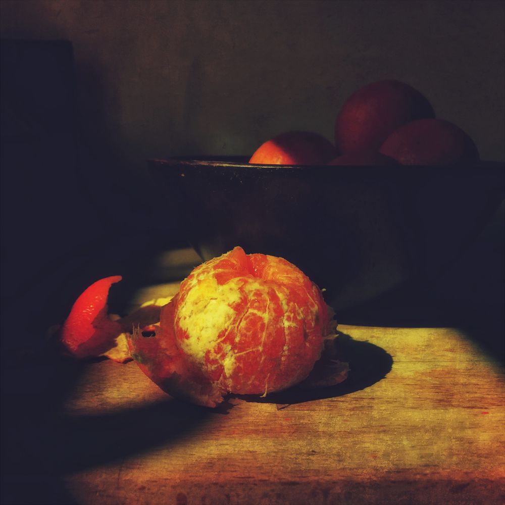 A peeled tangerine on a wooden surface in muted sunlight. there is a bowl behind with more fruit in.