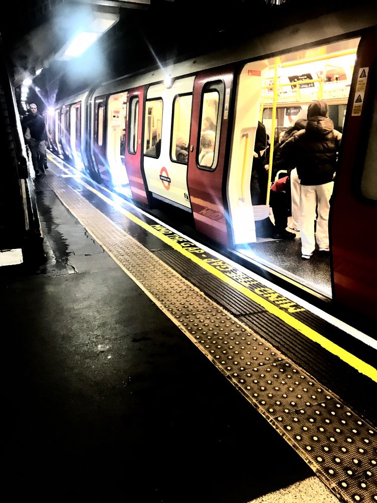 Moody lit underground image. There is a tube train and people. The platform is visible too with markings for safety.