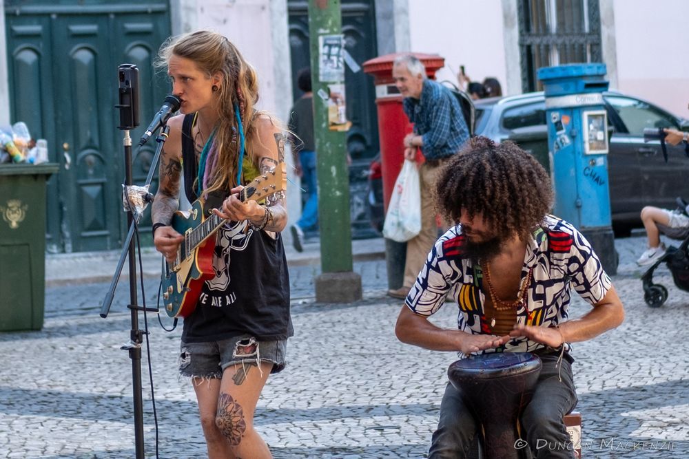 two musicians performing in an open plaza (one singing with a guitar, another playing a drum), man walking behind with his groceries