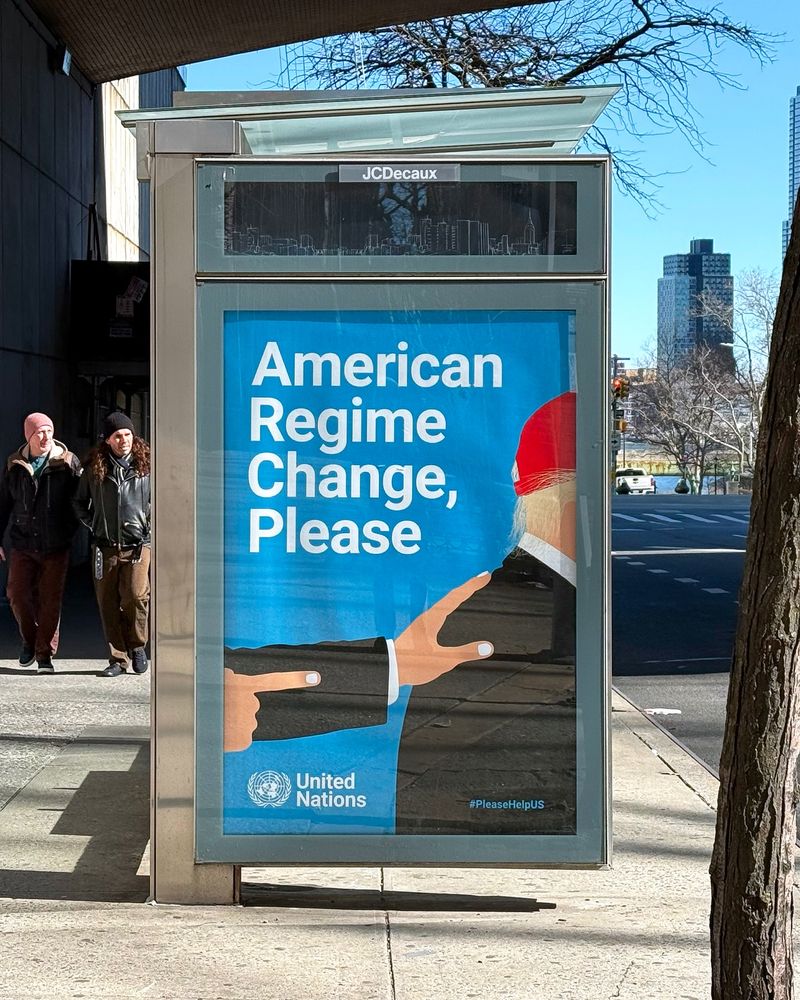 An official United Nations billboard advertisement that says "American Regime Change, Please" located at a bus stop next to the UN headquarters in New York City. Included on the ad is an image of Trump being pushed aside.
