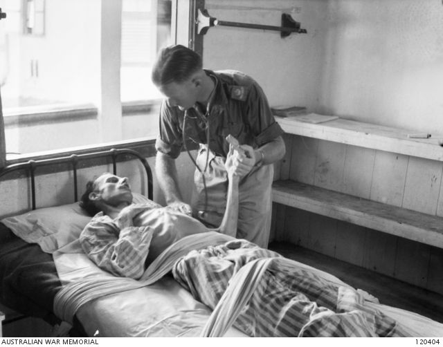 A black and white image of an emaciated man in striped pyjamas lying on an iron-framed bed in a bare room. His shirt is open and all his ribs are clearly visible, over a heavily sunken abdomen. A doctor in army fatigues, with his sleeves rolled up and a stethoscope around his neck, is holding the man's hand and placing a hand on his shoulder. The two men look at each with intense focus.