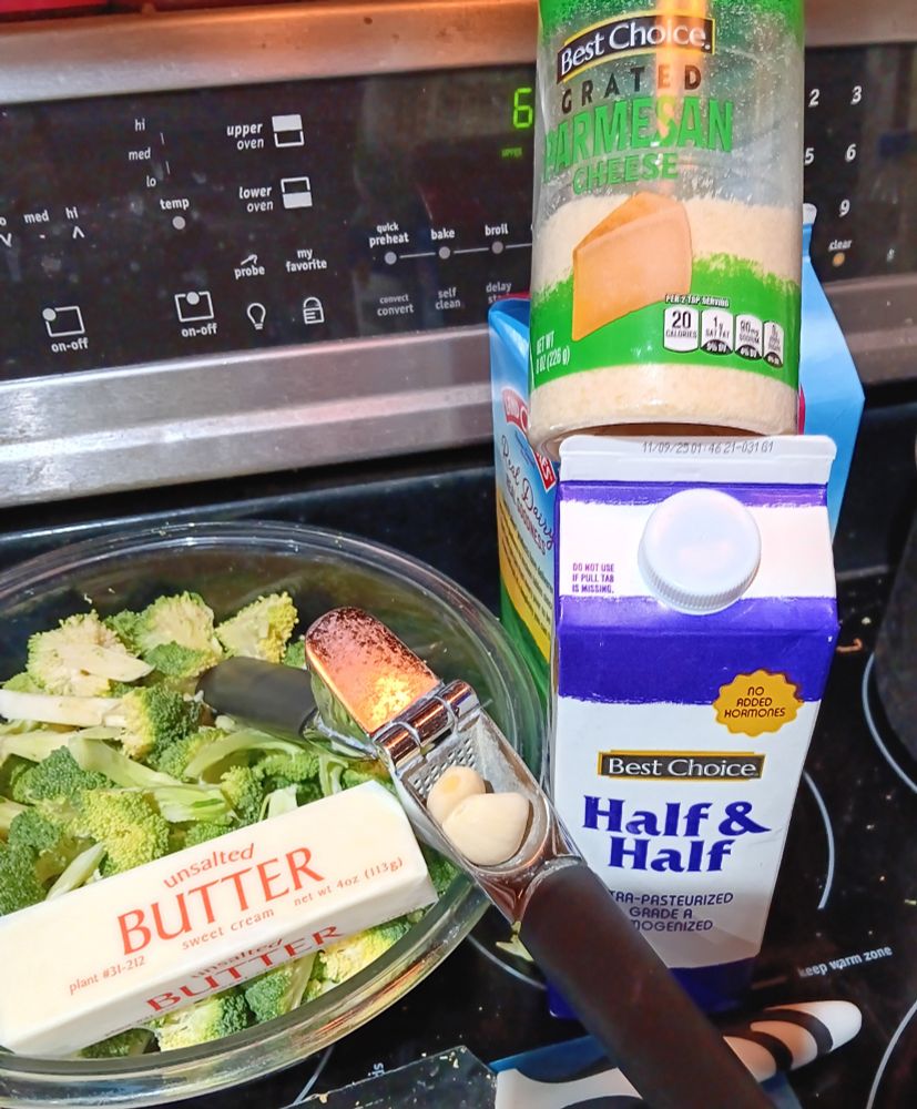 A bunch of ingredients gathered on the stovetop. A full stick of unsalted butter still in its packaging and two cloves of garlic in a garlic press sit in the bowl of chopped broccoli next to a couple of cartons of half & half with a container of grated parmesean resting on top of them.