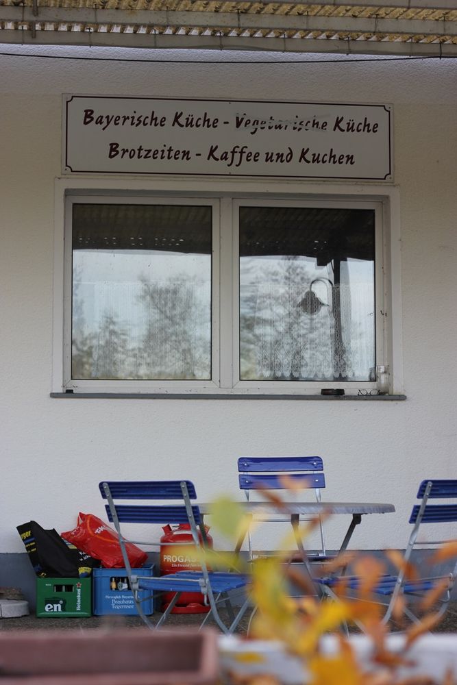 Eine Sitzgruppe (runder Tisch und vier blaue Biergartenstühle). 
Im Vordergrund ragt eine Pflanze unscharf in den unteren Teil des Bildes.

Hinter den Stühlen sieht man eine Hauswand mit einem Fenster. Über dem Fenster hängt ein Schild, auf dem geschrieben steht: "Bayerische Küche - Vegetarische Küche - Brotzeiten - Kaffee und Kuchen" wobei das Wort "Vegetarische" mit einem über Kreuz durchgestrichen ist.

Unter dem Fenster stehen eine rote Gasflasche und zwei Kästen Bier.