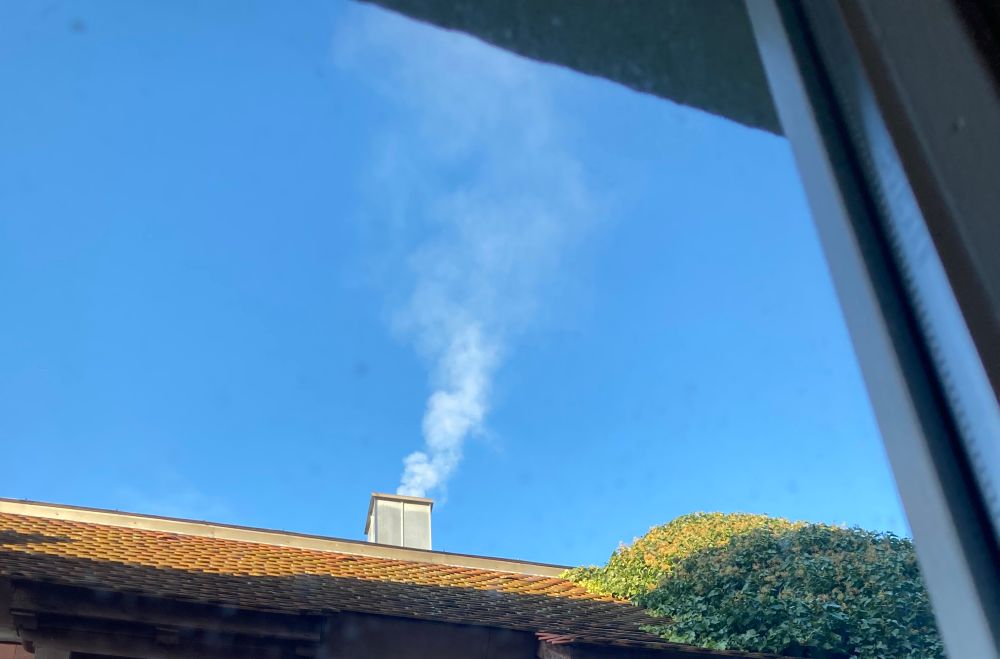 Aus einem Fenster nach draußen fotografiert sieht man einen Schornstein aus dem Rauch austritt