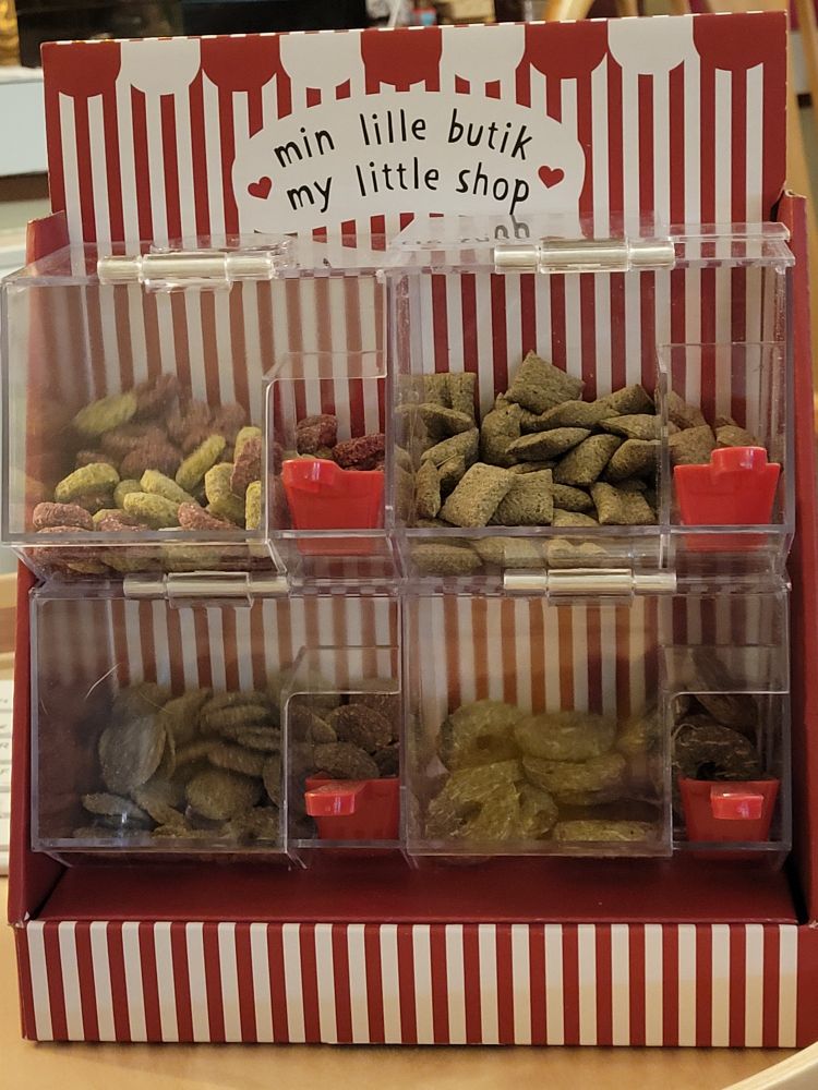 There is a set of transparent, plastic boxes or bins. There are two boxes stacked on two boxes. They are filled with different types of rabbit treats. The boxes are sat in a red and white striped holder. A sign on the holder reads "My little shop".
