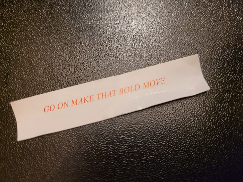 A white slip of paper with red writing which reads "Go on make that bold move". The background is shiny black with a bit of light reflecting in the top, middle of the photograph. 