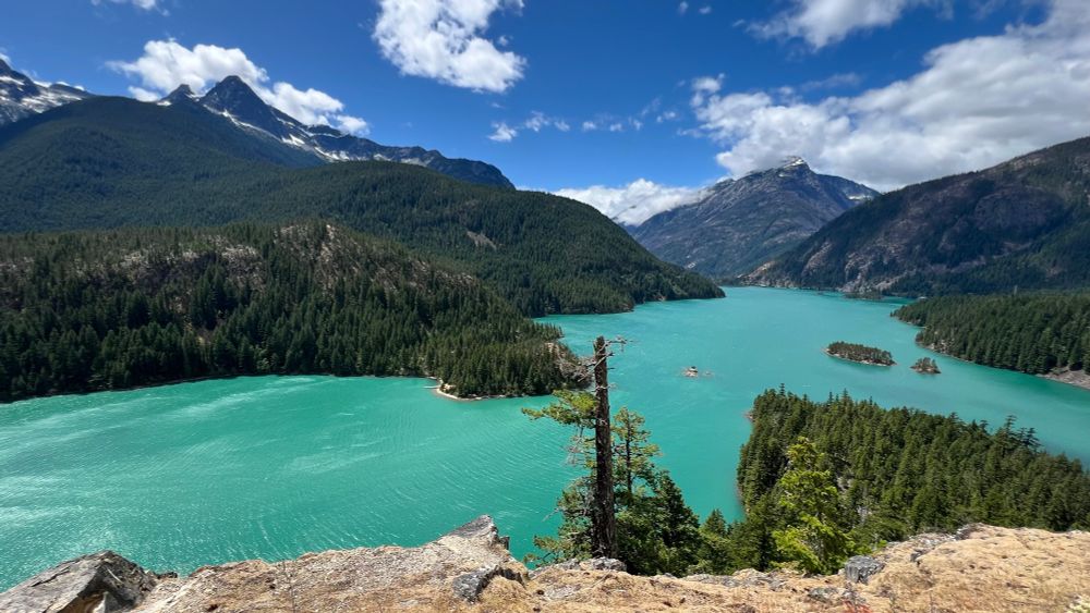Looking west across Diablo Lake, some evergreen trees in the foreground, mountains with small patches of snow in the background, and beautiful aquamarine-colored water that looks slightly milky due to “glacial flour,” which is very fine rock sediment suspended in the water. The sky above is deep blue with scattered white clouds.