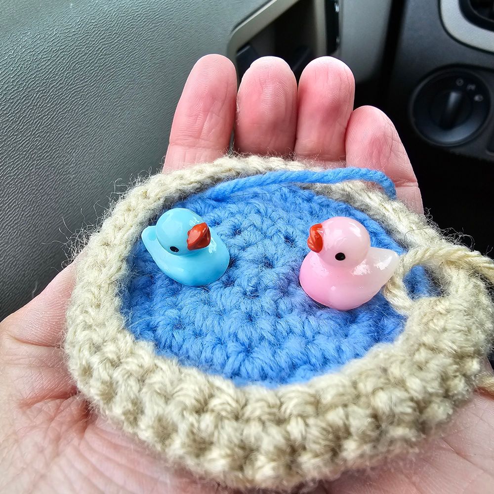 A hand cupping small fiber arts of a crocheted pond. On top of the pond, sit two little plastic ducks facing one another. 