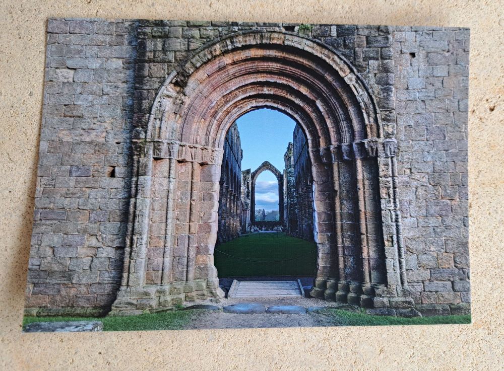 Postcard featuring a photo by Jay Hulme. An arched  portal in a gray stone wall gives a glimpse into a ruined church, with an empty window at the far end framing blue sky.