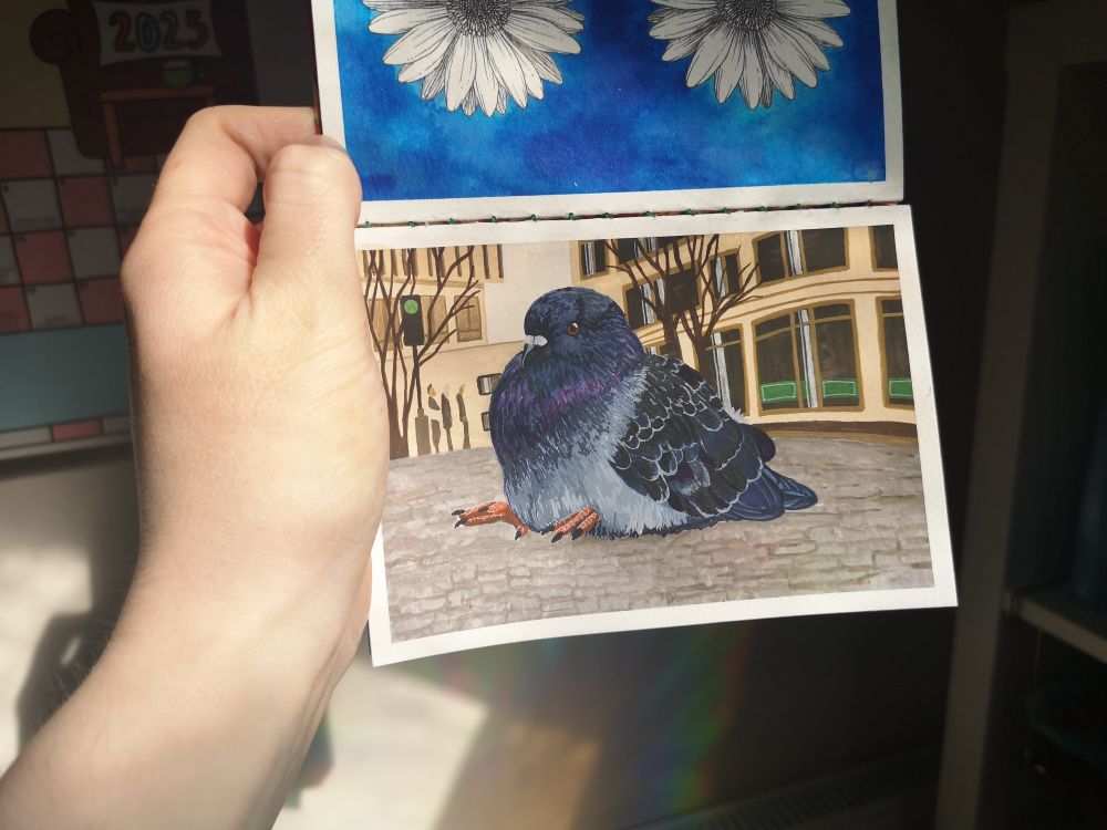 A hand from the left of the screen holds an open sketchbook page showing a finished gouache painting of a fat pigeon sitting on a cobbled street. His feet are poking out to the left. His feathers are grey and black with purple and teal highlights around his chest and neck. The background is a city street in shades of brown. There is light shining onto the sketchbook and the background of the photo is in darkness.