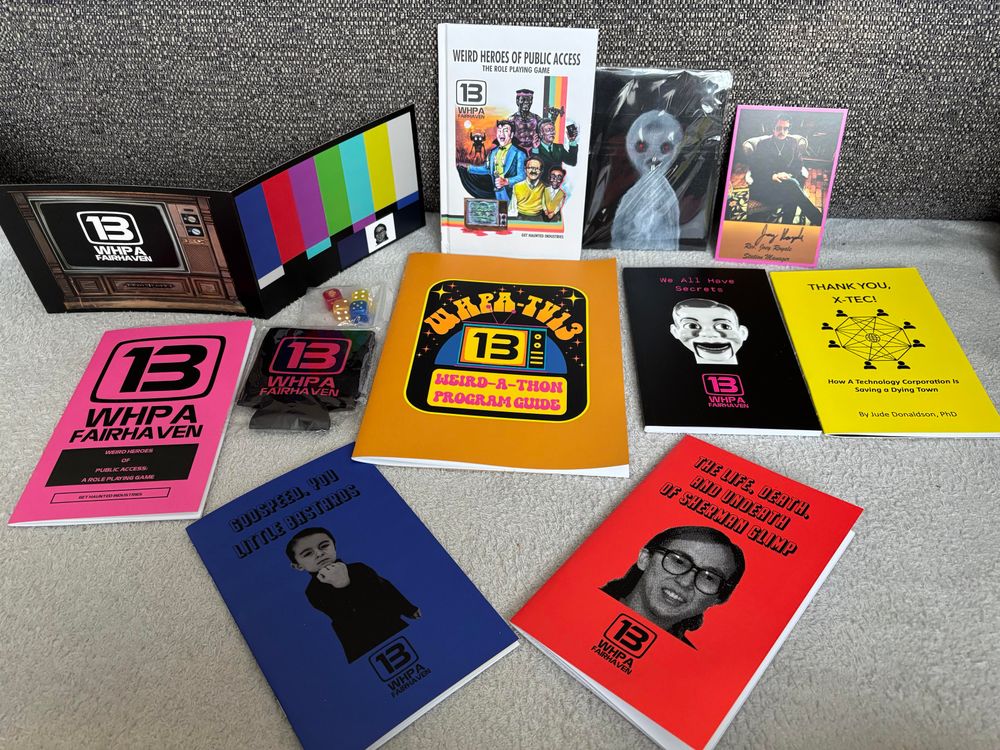 Various items for the Weird Heroes of Public Access RPG by Get Haunted Industries. 

Back row, left to right: miniature three fold GM screen, digest-sized hardcover rulebook, a small painting of a ghostly figure, and a signed postcard of the author.

Middle row: WHPA 13 Fairhaven zine, station logo can koozy, packet of four six-sided dice with thematic designs, Weird-A-Thon Program Guide, We All Have Secrets zine, Thank You X-Tec Zine.

Front row: Godspeed You Little Bastards zine and The Life, Death, and Undeath of Sherman Glimp zine.