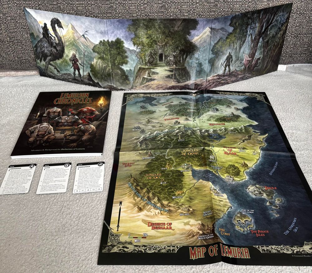 To the rear stands a trifold cardboard game master screen for the Barbarians of Lemuria RPG. It bears artwork of armored heroes approaching an ominous door. To the left is the Lemurian Chronicles book of adventures, bearing artwork of monsters with a single large eye in their chest. To the right is a poster map of the lands of Lemuria. To the front left are three printed cards for use with the Ironsworn RPG.
