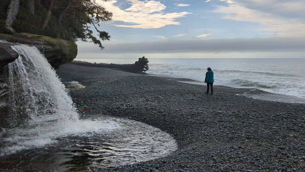 A waterfall over a sandstone ledge onto a beach with a person walking towards a log.