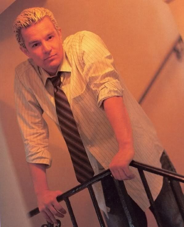 The image features a person with short, light-colored hair leaning casually on a black railing. They are positioned in a hallway with a warm, yellow-orange lighting that casts soft shadows. The person is wearing a light-colored, vertically striped shirt with the sleeves rolled up and a dark, diagonally striped tie that is loosely knotted. The background shows a blurred view of the hallway, implying depth and perspective, with no distinct objects in sight.